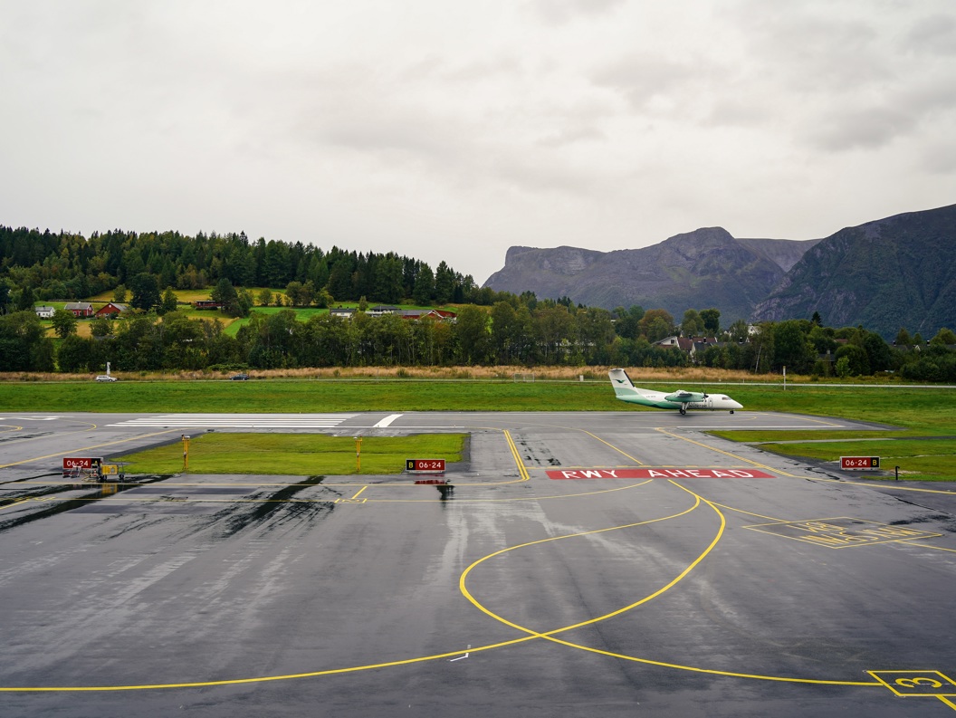 Flyplasslandskap med et lite passasjerfly på rullebanen, omgitt av grønne åser og fjell i bakgrunnen under en overskyet himmel.