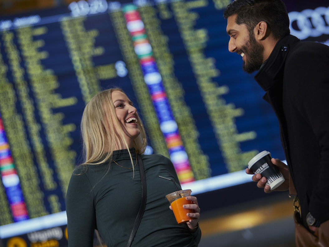 To personer ler og holder drinker foran en stor avgangstavle på flyplassen som viser flyinformasjon