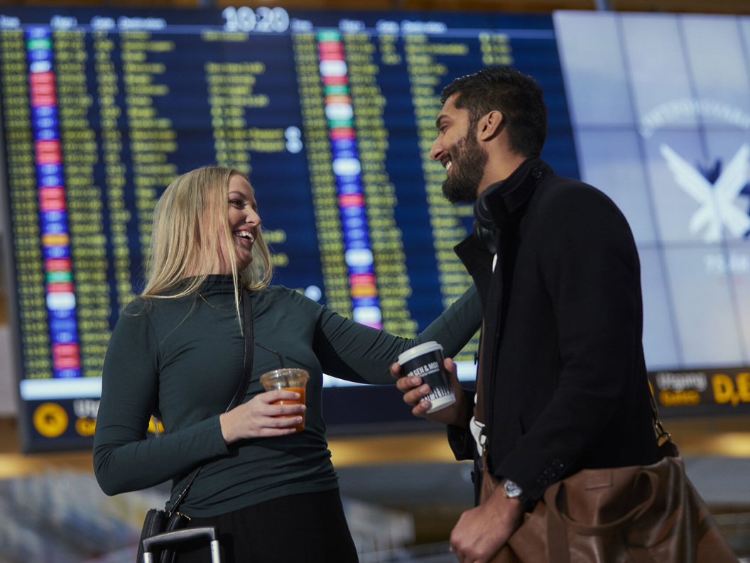 To personer smiler og holder drikke foran en flyplasstavle med flyinformasjon.