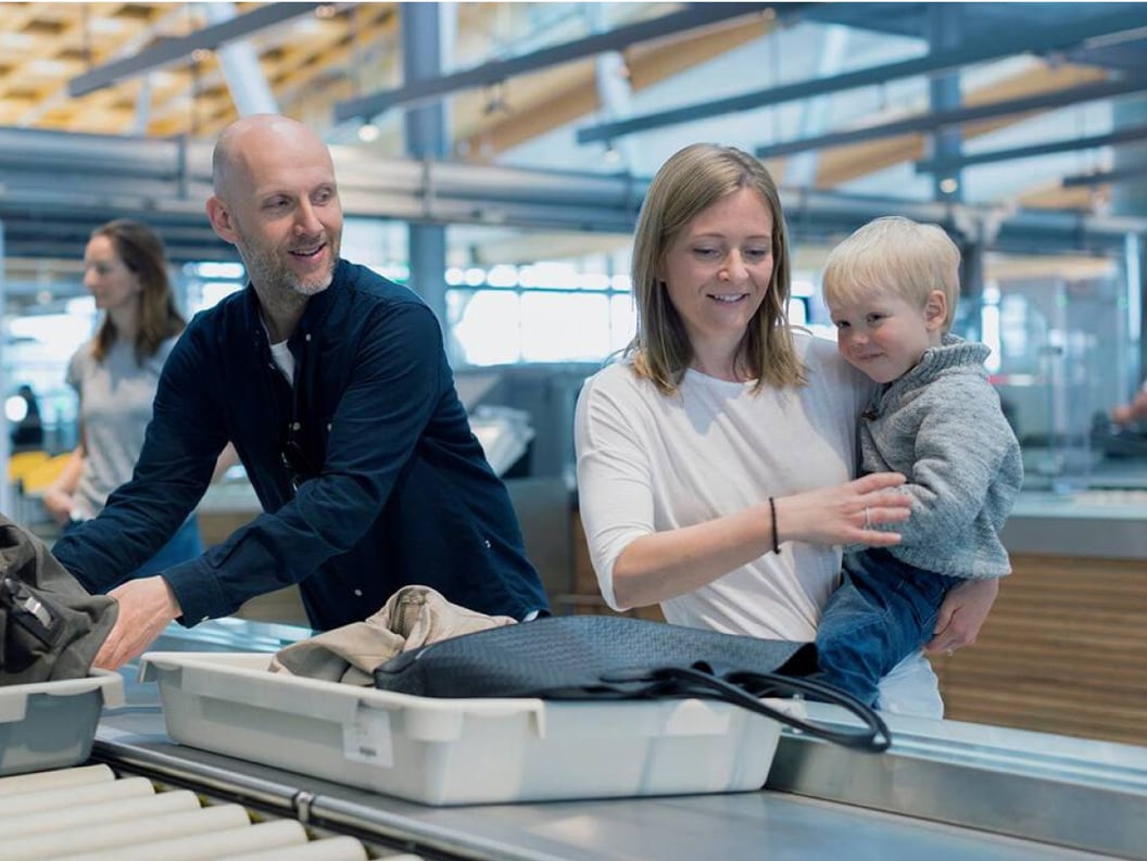 En mann og en dame som holder et lite barn står i sikkerhetskontrollen og legger tingene sine i grå esker som sendes avgårde. De er glade og smiler til hverandre.