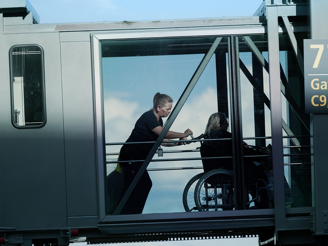 Dame som assisterer en annen dame i rullestol ut av flyet. Vi ser de gjennom vindusruten i overgangen mellom flyet og terminalen.