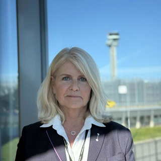 Kvinne i dressjakke med hvit skjorte står foran et glassvindu med utsikt over en flyplassterminal og kontrolltårn i bakgrunnen.