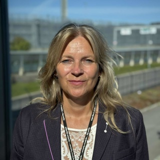 Kvinne i formelt antrekk smiler utendørs med skyfritt vær og bygninger i bakgrunnen.