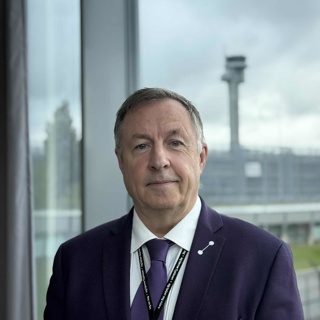 En mann i dress står foran et stort vindu med utsikt mot en flyplassterminal og kontrolltårn i bakgrunnen.