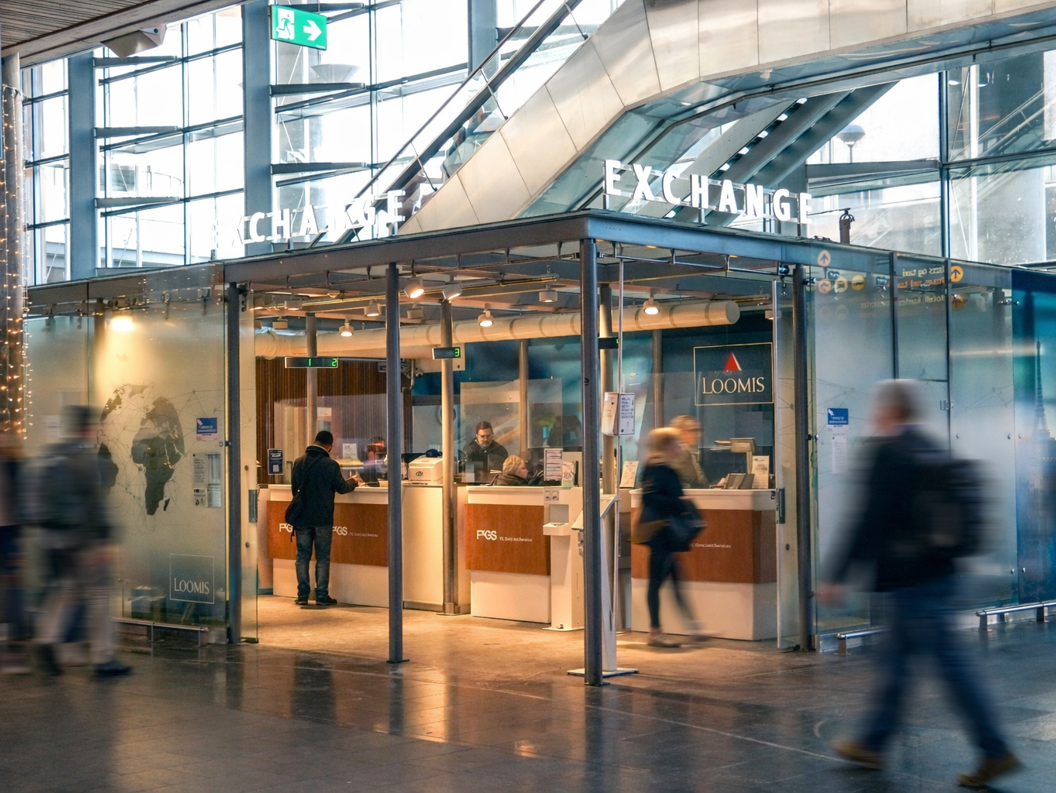 En person som bruker en minibank for utenlandsk valuta på en flyplass, med flere valutamuligheter vist på en skjerm i nærheten, som indikerer internasjonal reise og finansielle transaksjoner