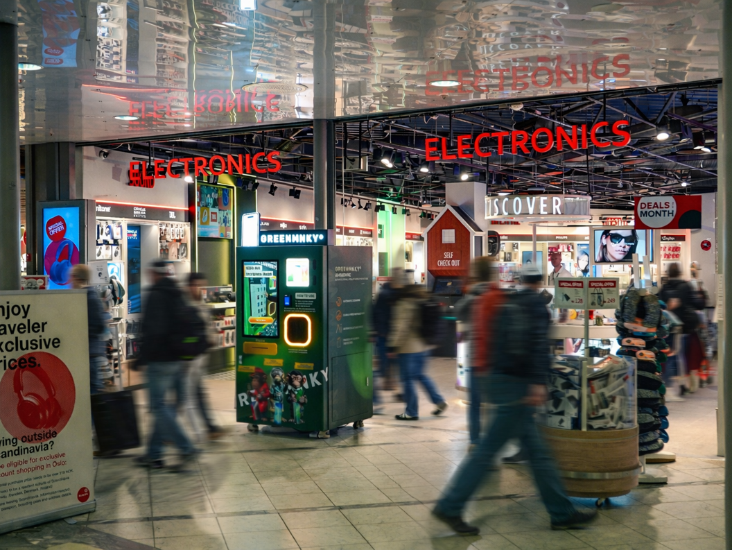 Store employee assisting a customer at a CAPI electronics retail display, featuring tech gadgets and accessories, in a brightly lit setting