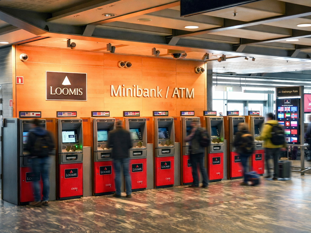 A person using a foreign currency ATM at an airport, with multiple currency options displayed on a screen nearby, indicating international travel and financial transactions