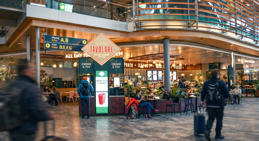 Baransatte som jobber bak en moderne bardisk, smilende og organiserer drikkevarer, med et stilfullt interiør med hengende lamper og ordkunst på veggene