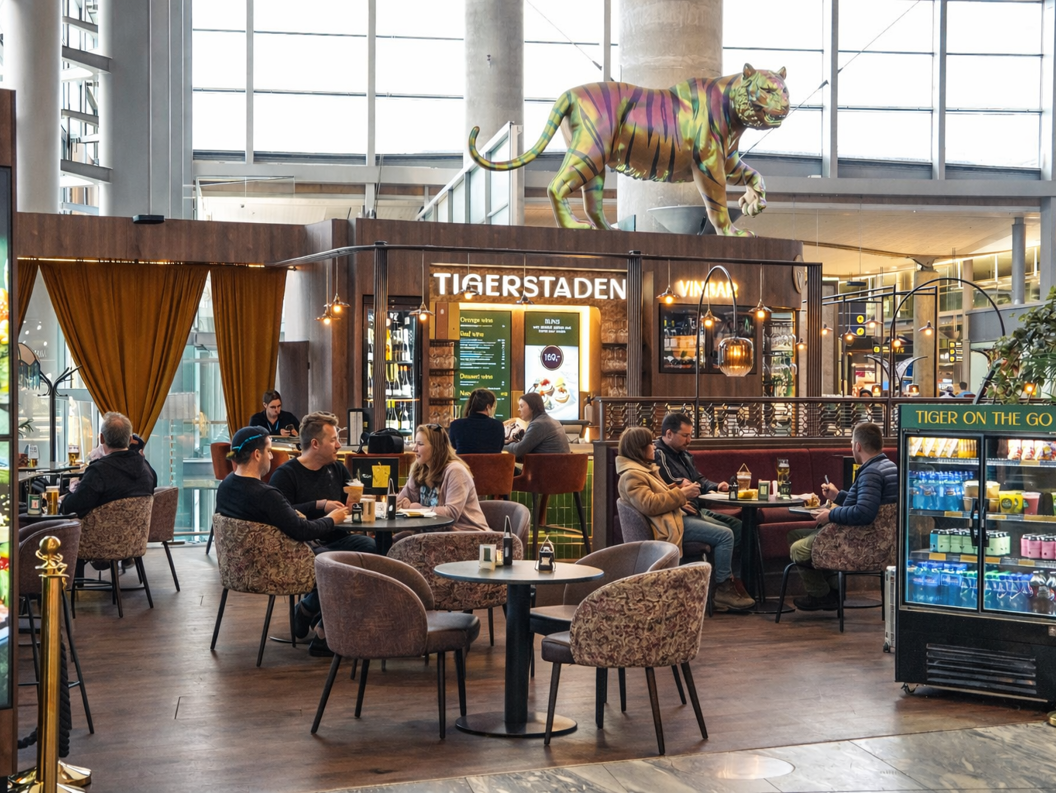 Moderne flyplassbar og loungeområde med sitteplasser, med en stor dekorativ tigerskulptur over "Tigerstaden Vinbar" skilt, med gjester og personale synlige, og et retningsskilt for porter E, F, G i bakgrunnen