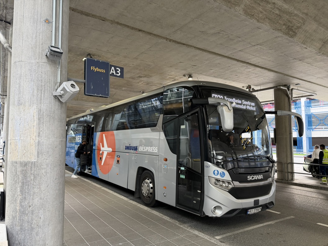 Blå buss utenfor en flyplassterminal i Norge, med passasjerer som stiger av og noen står med bagasje. Moderne bygning og parkeringsplass i bakgrunnen. Grå himmel og fuktig vær.