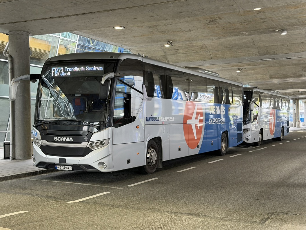To moderne Scania-busser parkert ved en terminal under et betongtak, med "Værnes Ekspressen"-merking, og destinasjonsskilt for Trondheim Sentrum, rute FB73