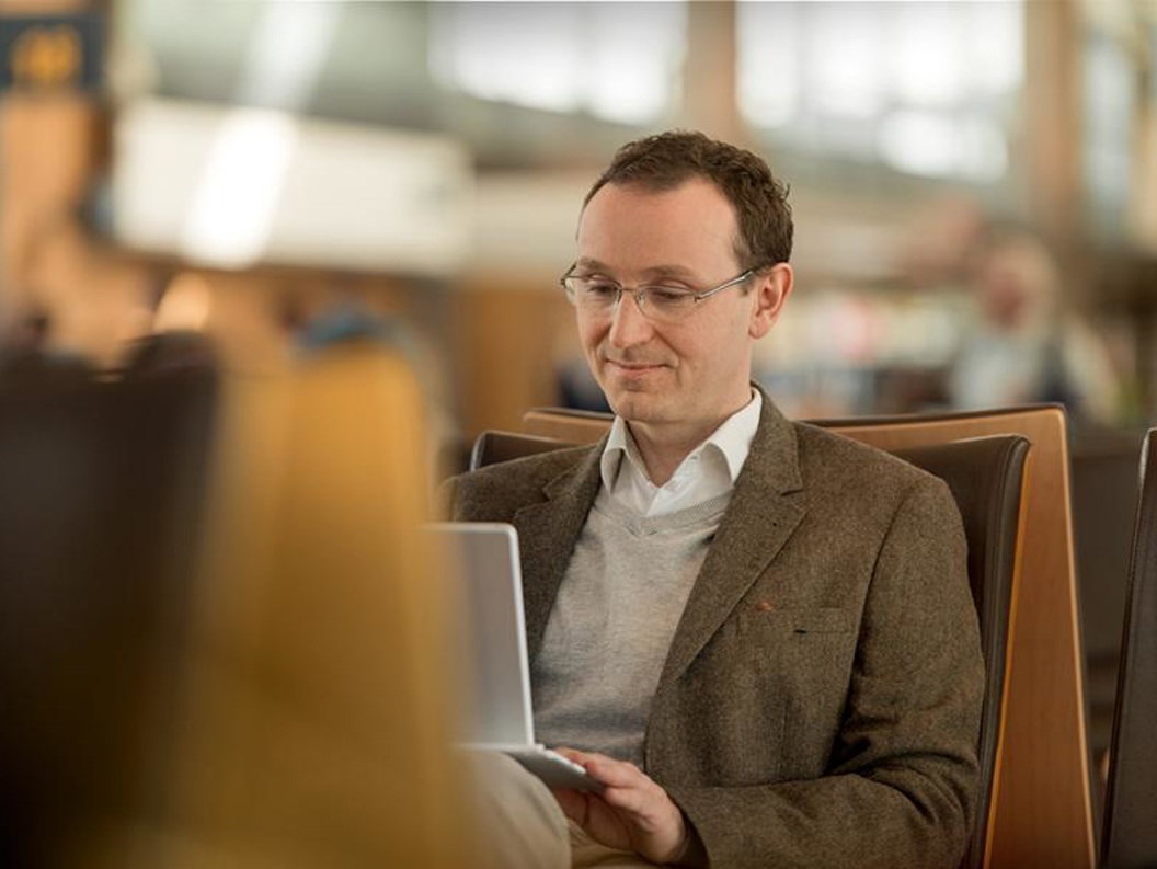 En mann med kort brunt hår og briller som sitter på en av plassene ved en gate og ser på sin bærbare pc som han har på fanget.